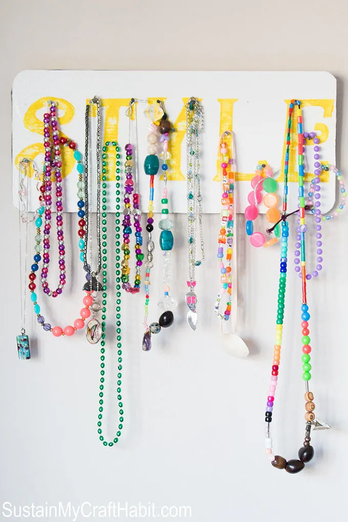 An accessory organizer holding colorful necklaces and bracelets. The word Style is stencilled on the jewelry display with yellow paint.