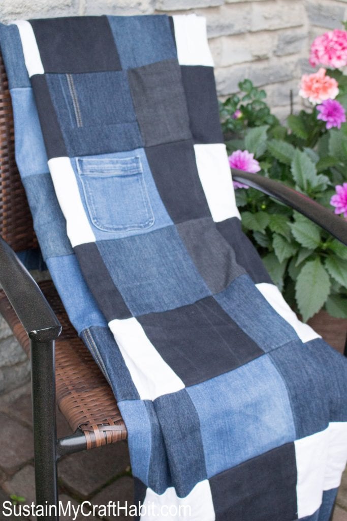 A checkered picnic blanket made from different colored denim, draped over a chair on a front porch. A basket of pink flowers are in the background.