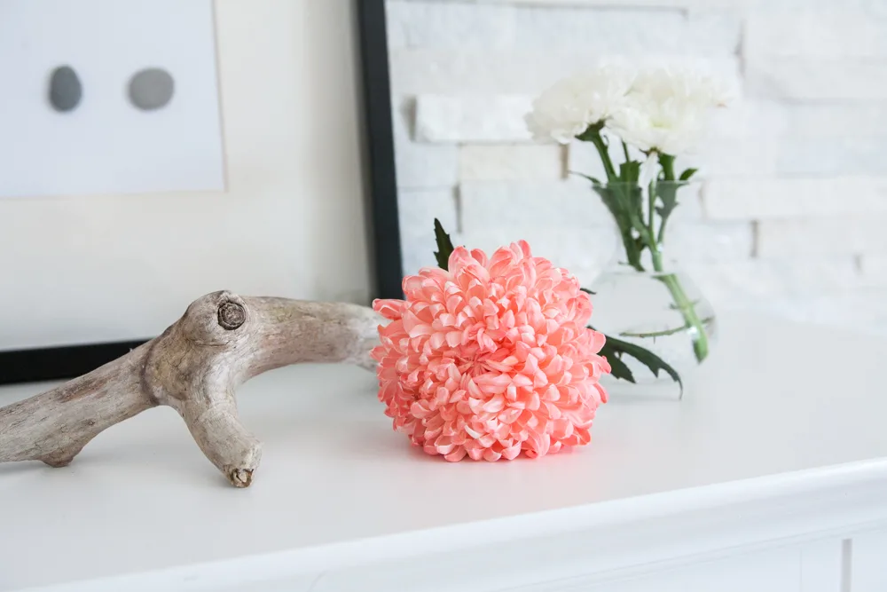 A coral chrysanthemum on a white fireplace mantel with a piece of driftwood and DIY wall decor in the background 