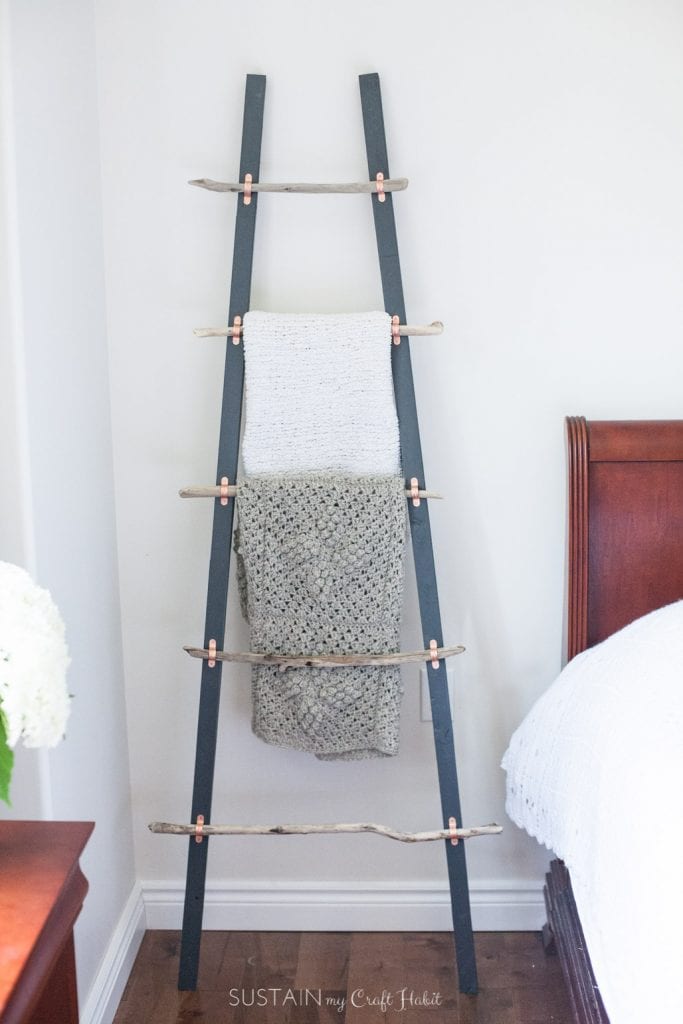 DIY driftwood blanket ladder leaning against a white wall in a bedroom.