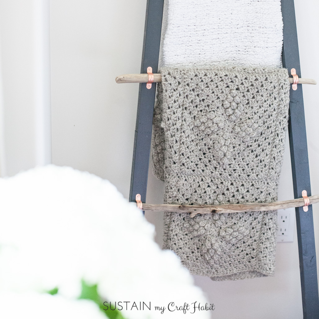 A white knitted and beige crocheted blanket hanging on the rungs of a driftwood blanket ladder.