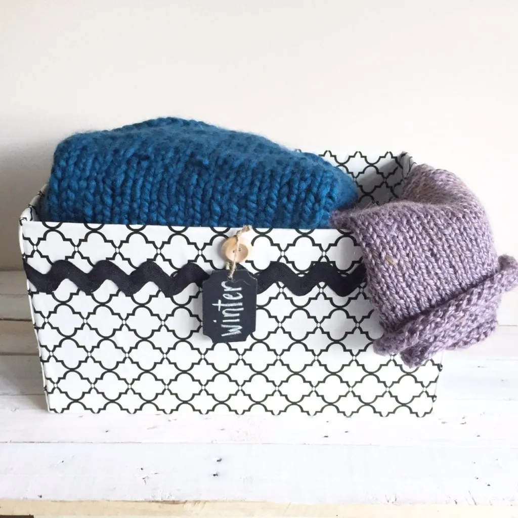 A completed decorative storage box filled with winter hats and scarves. A small chalkboard tag pinned to the front of the box reads winter.