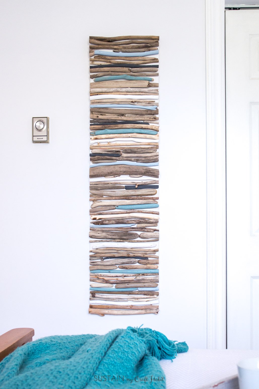 Completed painted driftwood art hanging on a white wall. A teal blanket is on a bed in the foreground.