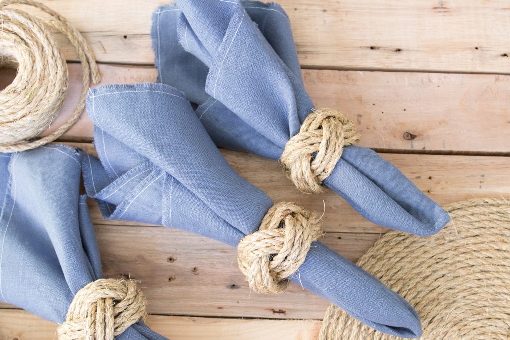 Three blue linen napkins on a rustic wood surface tied with DIY nautical knot napkin rings.