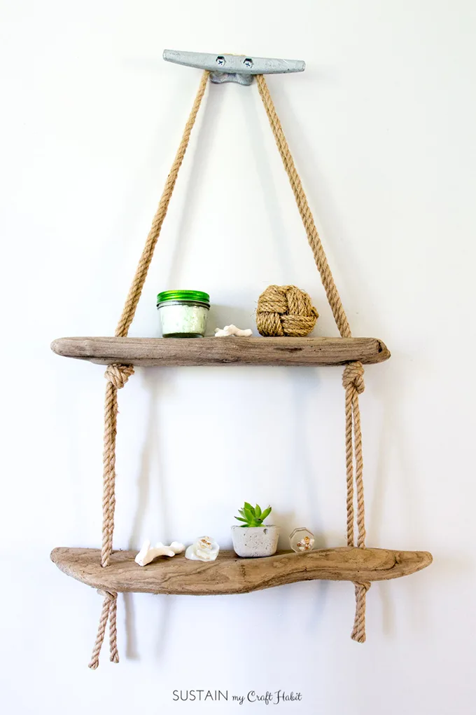 Two pieces of driftwood joined with rope and hanging from a decorative anvil on a white wall to form driftwood shelves.