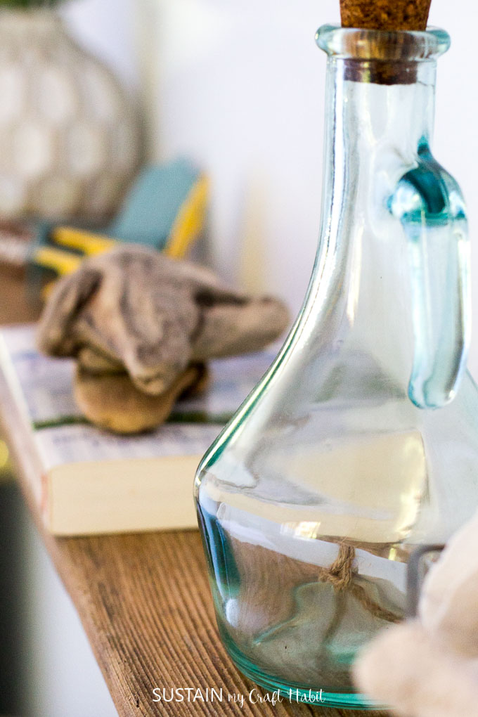 Close up view of a light teal glass bottle with a wrapped piece of paper inside, on a DIY driftwood shelf adorned with other beachy finds.