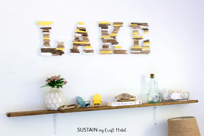 Rustic DIY shelf made with driftwood and inexpensive hardware store brackets mounted on a white wall. A sign saying LAKE is on the wall above the shelf.