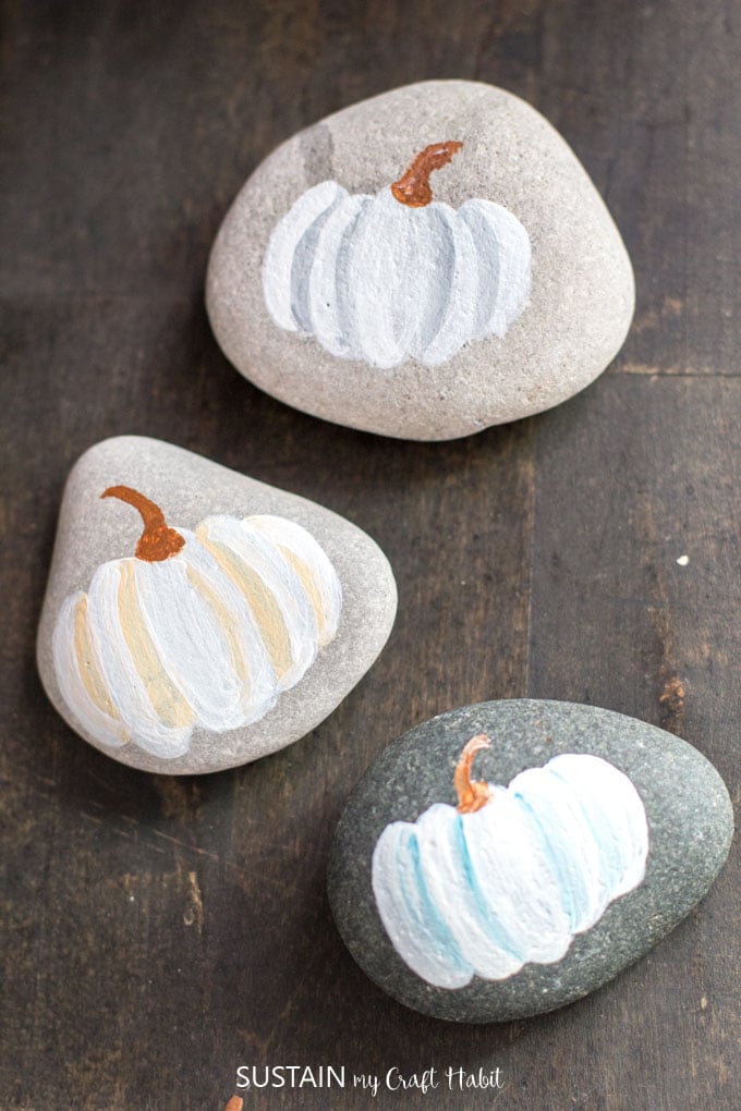 Three rocks painted with watercolor inspired pumpkins in gray, orange on teal. The rocks are resting on a dark wood surface.