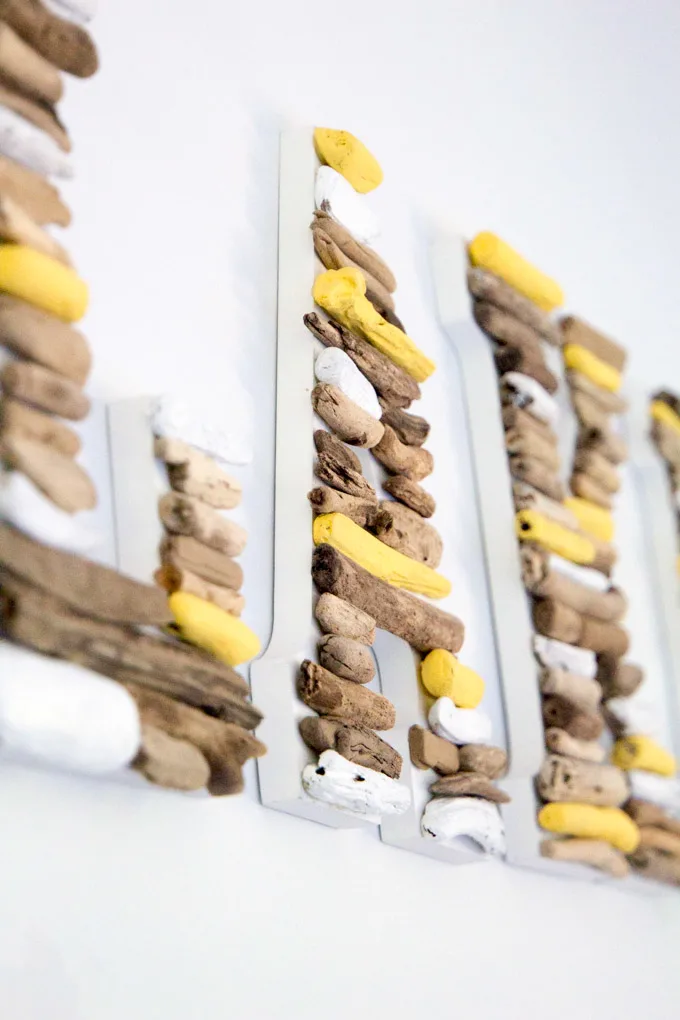 Close up image of the letter A made with pieces of driftwood painted yellow and white. The entire LAKE sign hangs on a white wall.
