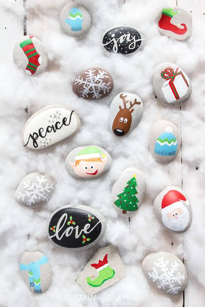 A collection of Christmas themed painted rocks including a reindeer, Santa, tree, snowflakes and more on a white wood surface.