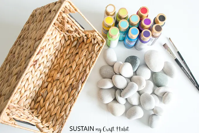 Top view of round beach stones and a variety of acrylic craft paints beside a basket on a white surface