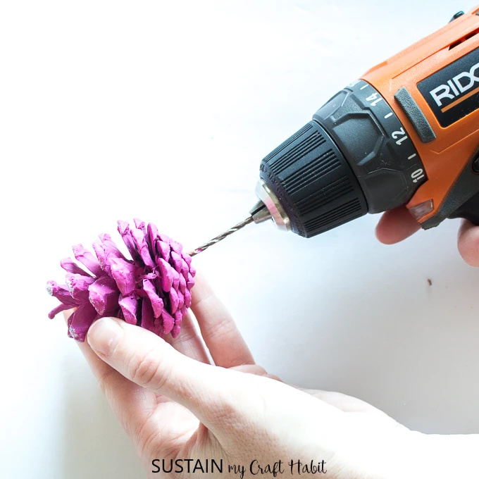 Drilling a hole at the bottom of a painted pine cone.