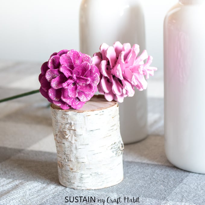 Pine cones painted in two shades of pink, with a dusting of glitter, resting on a small birch branch stump. 