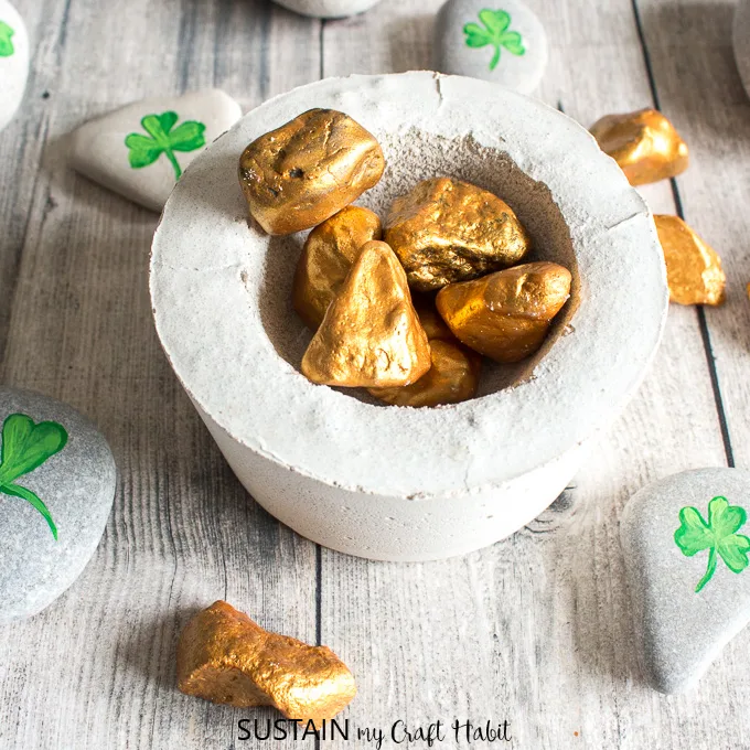 A small cement pot filled with gold painted rocks surrounded by rocks with shamrocks painted onto them.