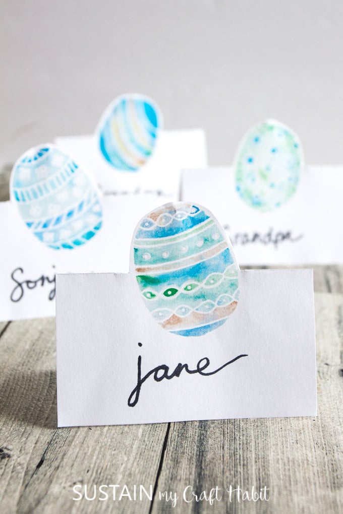 Four blue and green watercolor Easter egg place cards on a weathered wood surface.