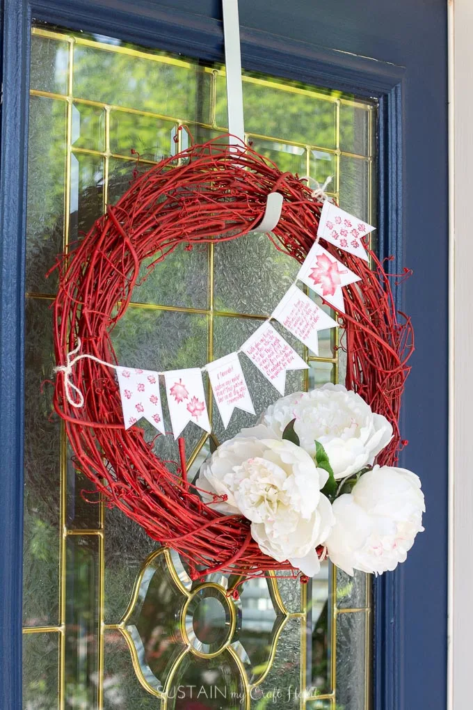 A red grapevine wreath decorated with white flowers and a small banner hanging on a blue painted front door
