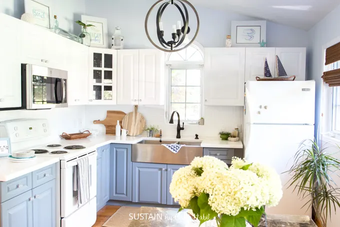 Entire view of a coastal style kitchen with white and gray painted cabinets decorated with touches of wood. A vase of white hydrangeas is in the foreground.