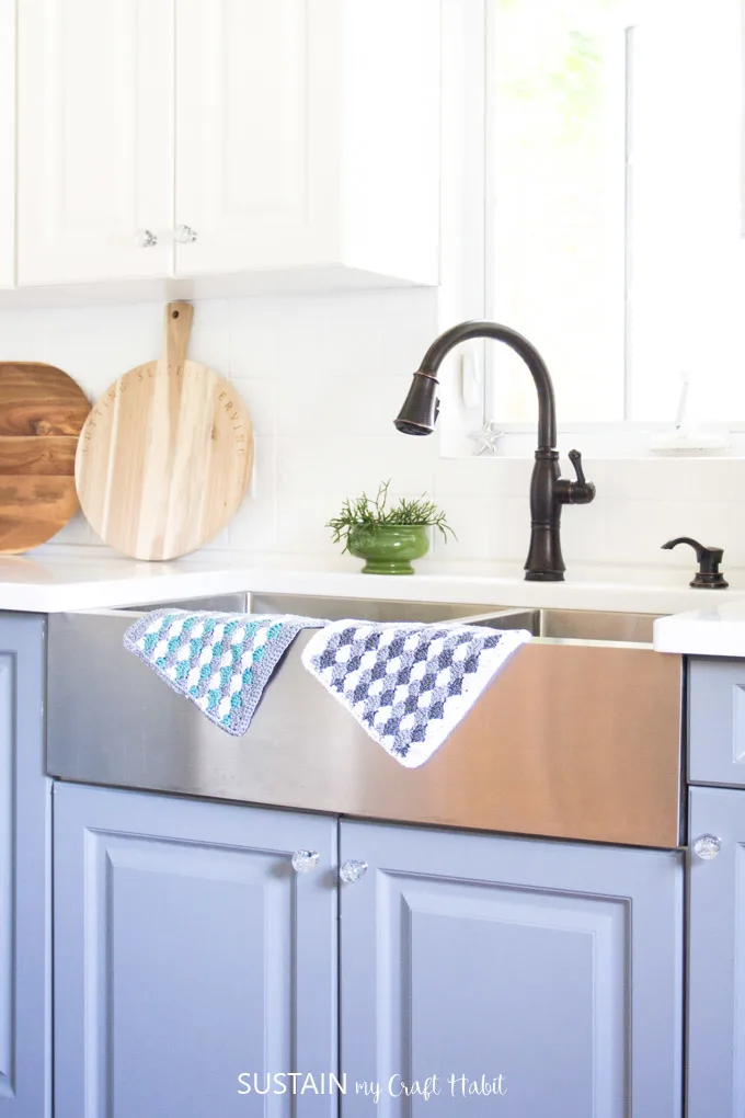 Two crochet dishcloths hanging over the edge of a farmhouse style sink.