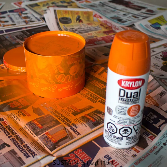 Spray painting an old cookie tin with orange Krylon paint on a newspaper covered surface