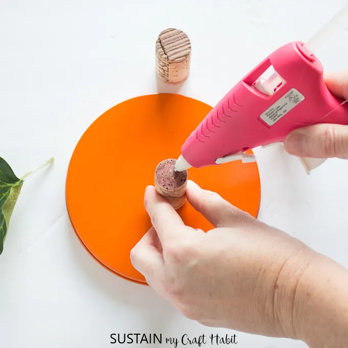 Using a hot glue gun to attach a wine cork to the top of a cookie tin lid painted orange