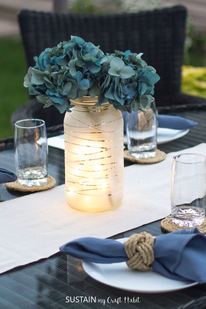 An outdoor table at dusk, set with dishes and lit by an upcycled pickling jar lantern with twinkle lights