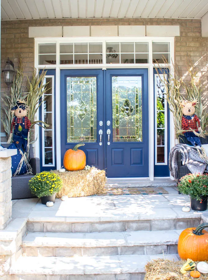 A fall front porch decorated with hay bales, orange pumpkins, mums, cornstalks and scarecrows