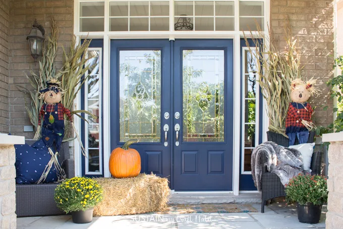 A front porch decorated for fall with orange pumpkins, hay, mums, scarecrows and cornstalks