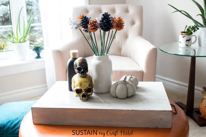 A vignette of orange, white and black pine cone flowers in a white glass vase surrounded by Halloween decorations