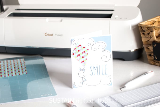 A handmade greeting card with a small kite and the word "smile". The card is propped up on a table in front of a Cricut Maker machine.