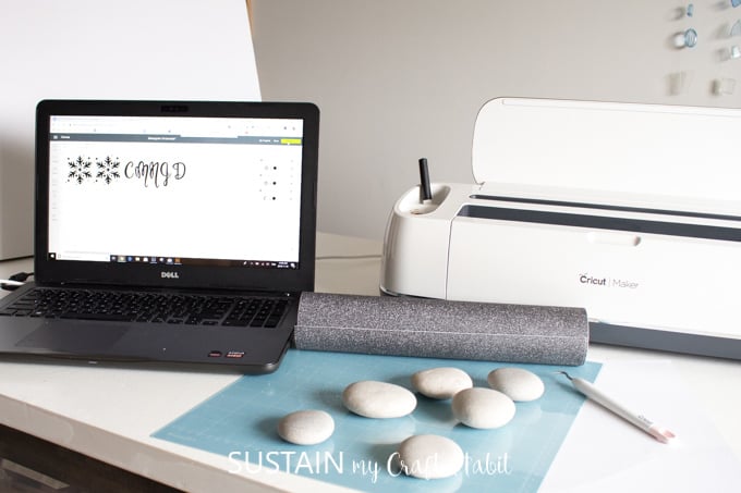 A laptop connected to a Cricut Maker machine displaying an image of the Cricut Design Space with a snowflake and monogram design loaded