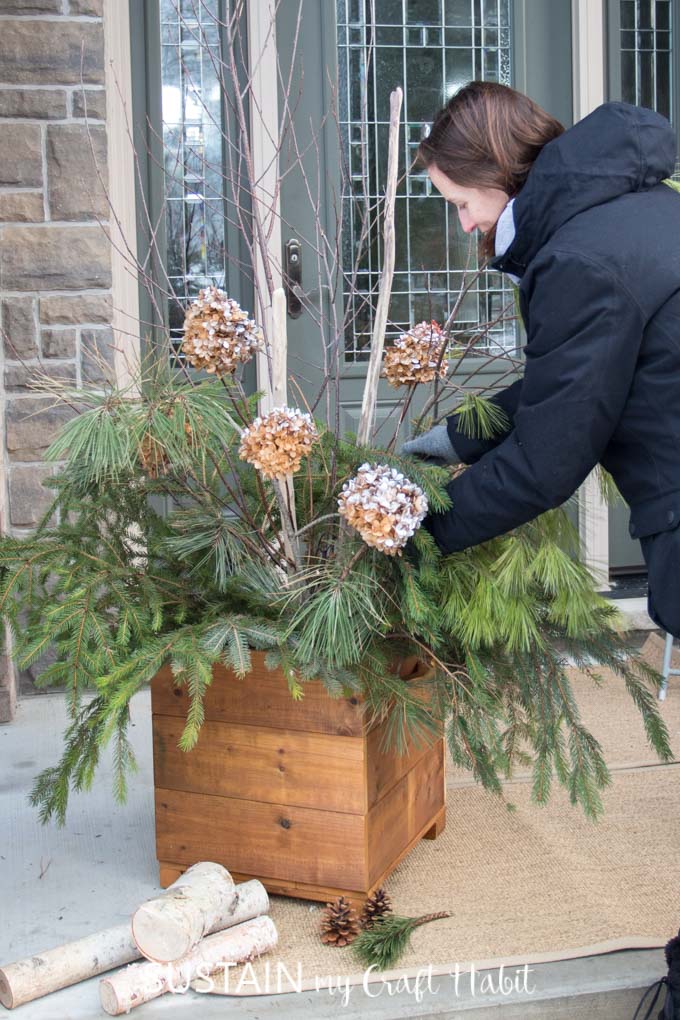 Woman filling an arrangement with branches and dried hydrangeas in a wooden DIY planter box