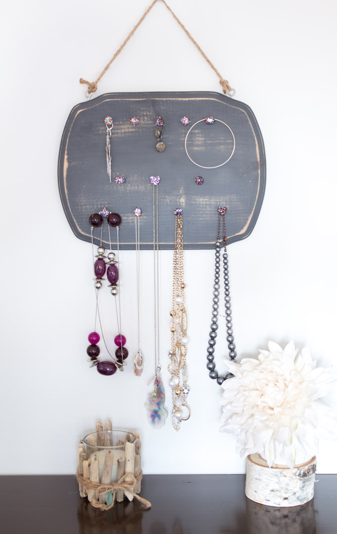 A dark grey wood plaque jewelry organizer hung on the wall with various pieces of jewelry hanging from it.