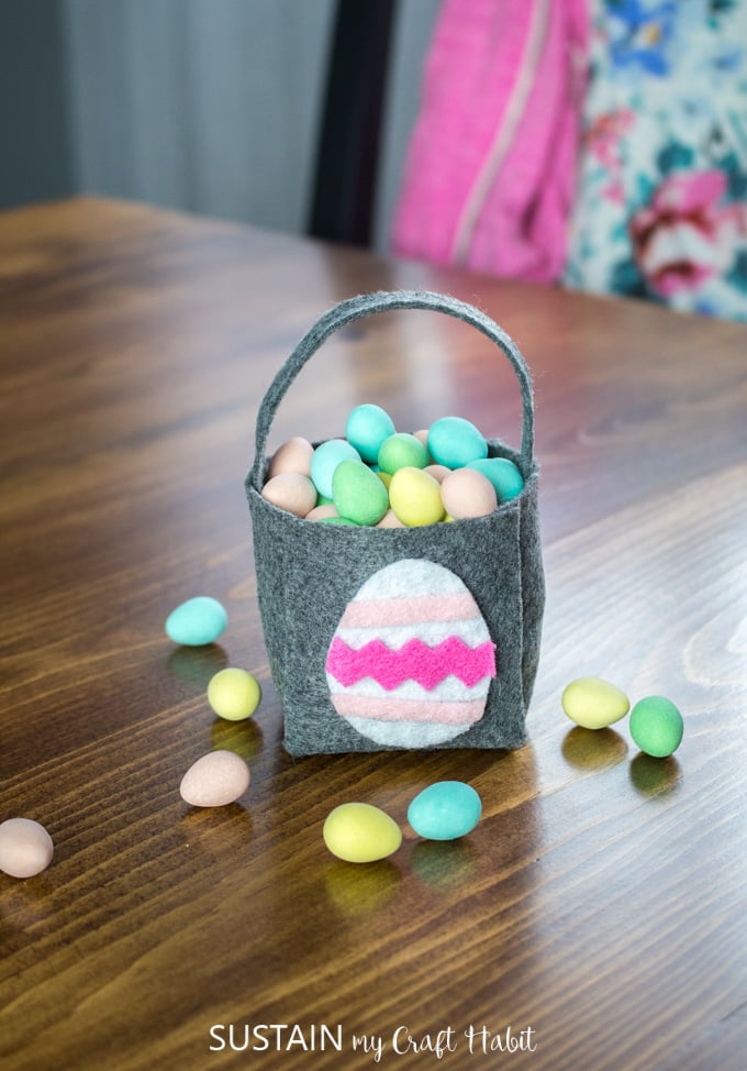 Gray felt Easter basket filled with miniature candy eggs on a dark wood table.