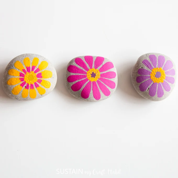 Yellow, pink and purple painted flower rocks side by side on a white background.