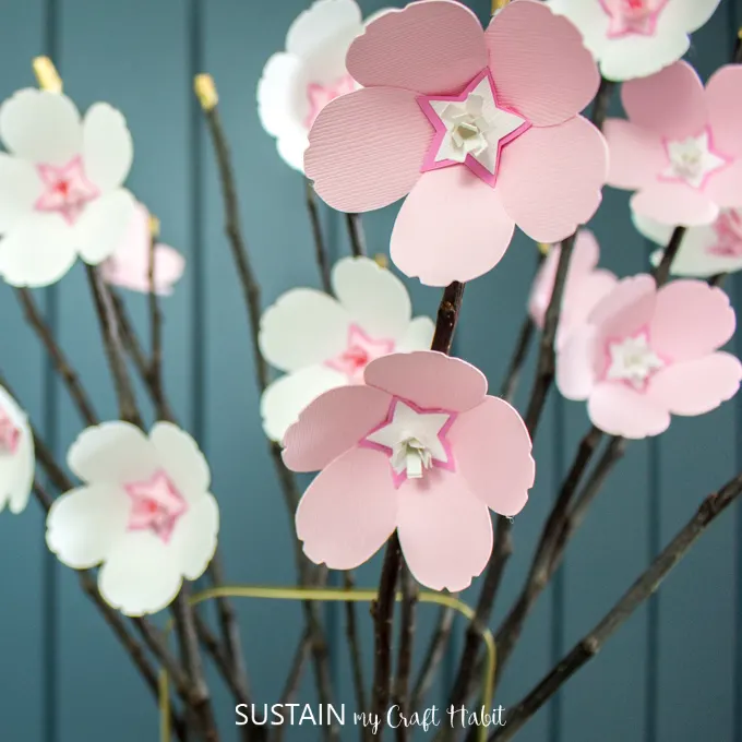 Pink and white paper cherry blossoms glued onto twigs against a teal blue wall