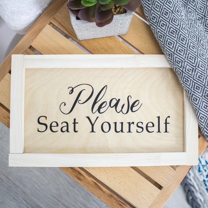A handmade wood sign on a wooden bench in a bathroom. The phrase "Please seat yourself" is written on the sign.