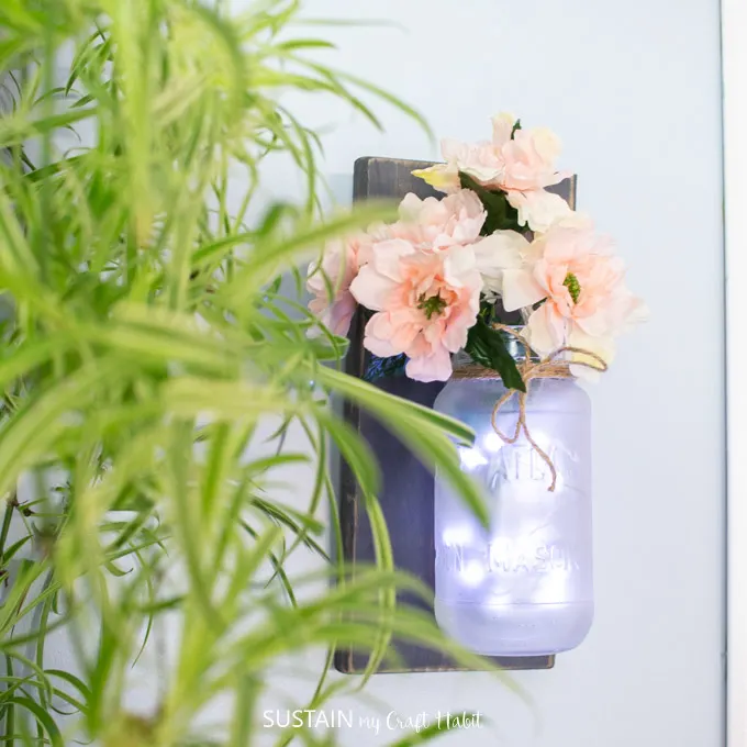 A frosted Mason Jar hanging from a wooden backing, filled with pink flowers and twinkle lights.