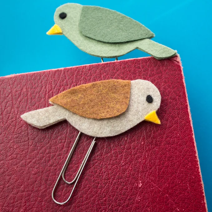 A brown felt bird bookmark lying on the surface of a red leather book cover. A second green felt bird bookmark is peaking out of the top of the book.
