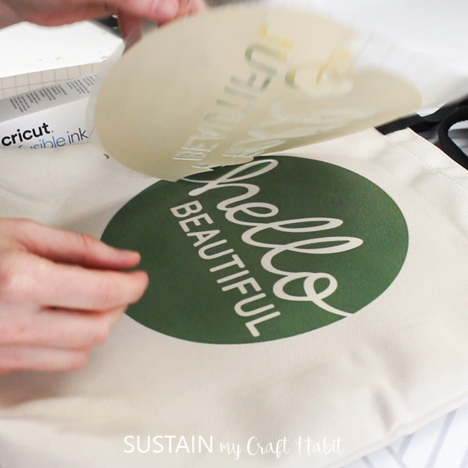 Lifting a sublimation transfer sheet off of a tote bag surface to reveal a sublimated design: a green circle with the phrase hello beautiful inside.
