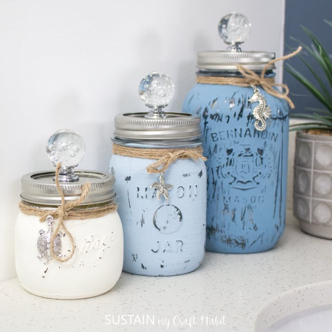 Three painted mason jars in different shades of blue, styled on a white bathroom counter.