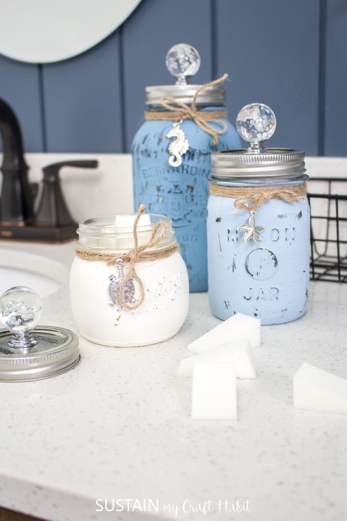Three painted mason jars in various shades of blue sitting on a white bathroom counter. The jars are distressed and embellished with coastal style charms and a glass knob.