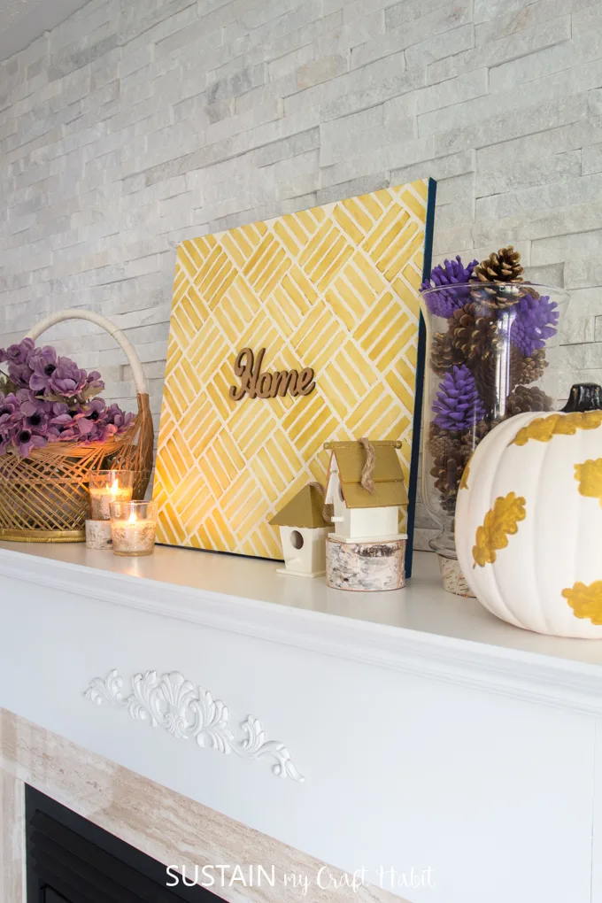 A fireplace mantel decorated for the fall with elements in gold, lavender and cream. Includes a large painted canvas, basket filled with mauve anemone flowers, vase of lavender pine cones and more.