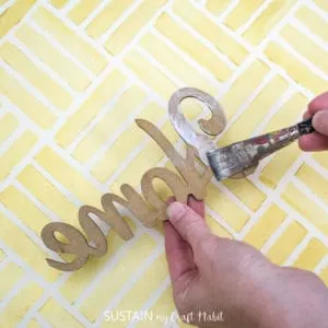 woman using paint brush to put glue on the back of a wooden cut out that spells "home".