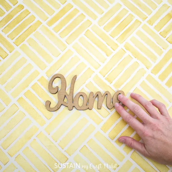 woman glueing wooden cut out of the word "home" onto completed stencilled canvas