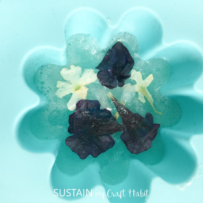 Dried flowers being pressed into melted soap base in a flower-shaped silicone mold.