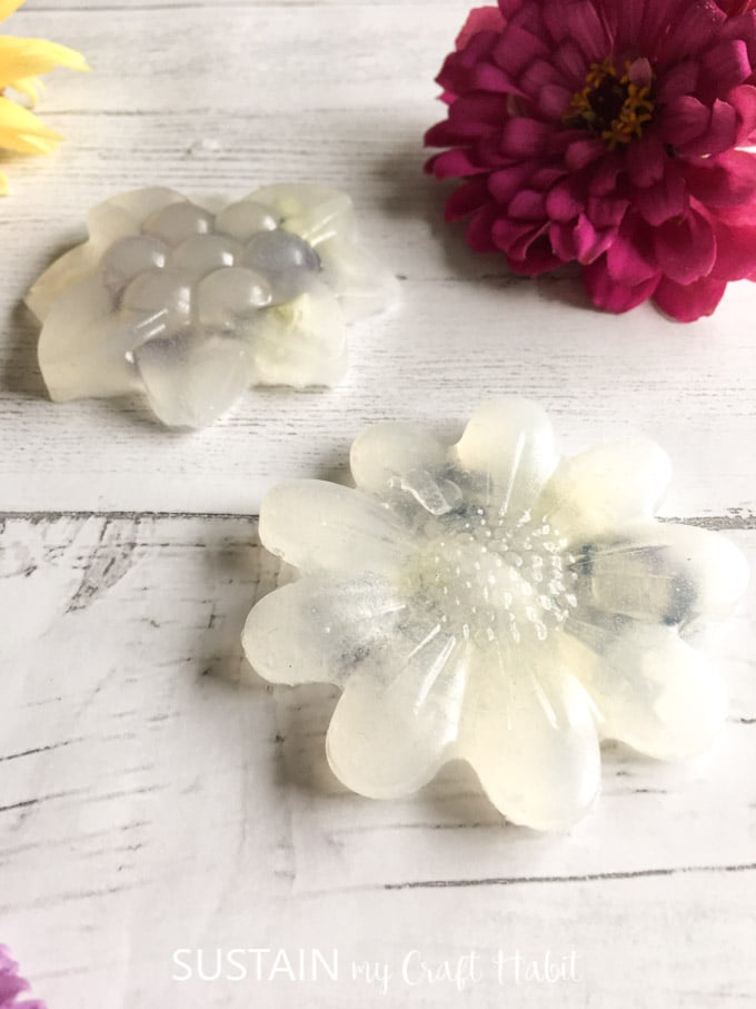 Two translucent glycerin soaps on a white wood plank surface with a dark pink zinnia flower in the background.