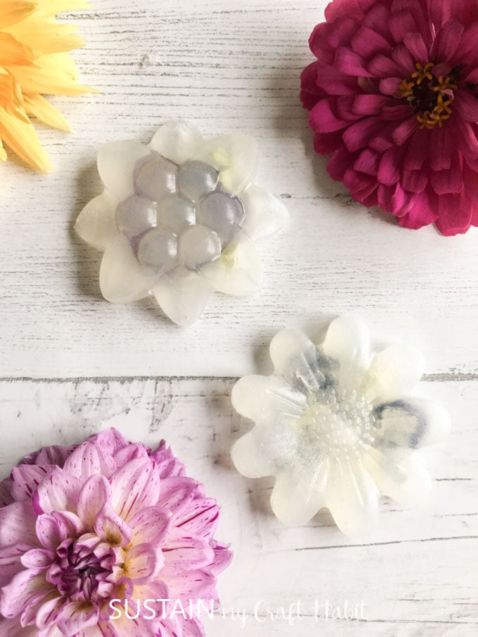 Overhead image of the two completed glycerin soaps on a white wood background, surrounded by pink, purple and yellow fresh flowers.