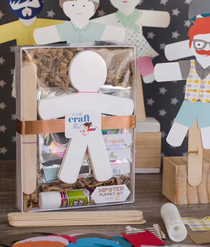 Paper dolls, glue, wooden sticks, and paper accessories wrapped in a box placed beside finished paper dolls on wood stands.