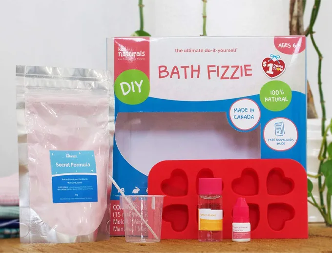 Ingredients needed to make a bath bomb. The kit includes a bag with the words "secret formula", a red silicon mold, cup and stir stick and oils.
