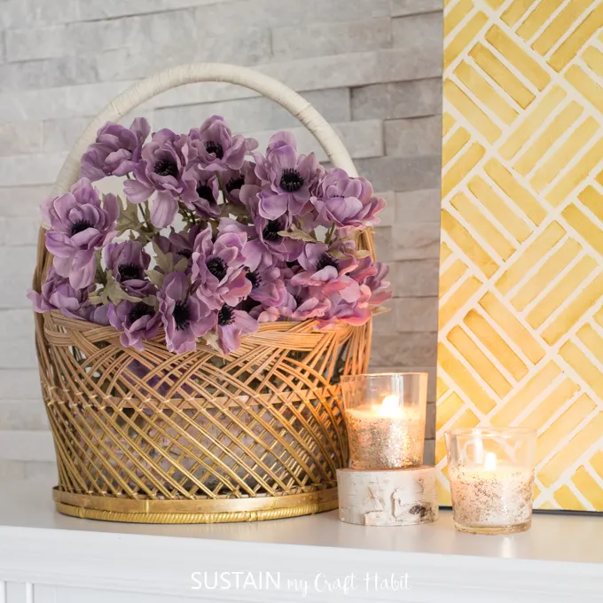Gold painted wicker basket with purple flowers beside 2 lit candles.